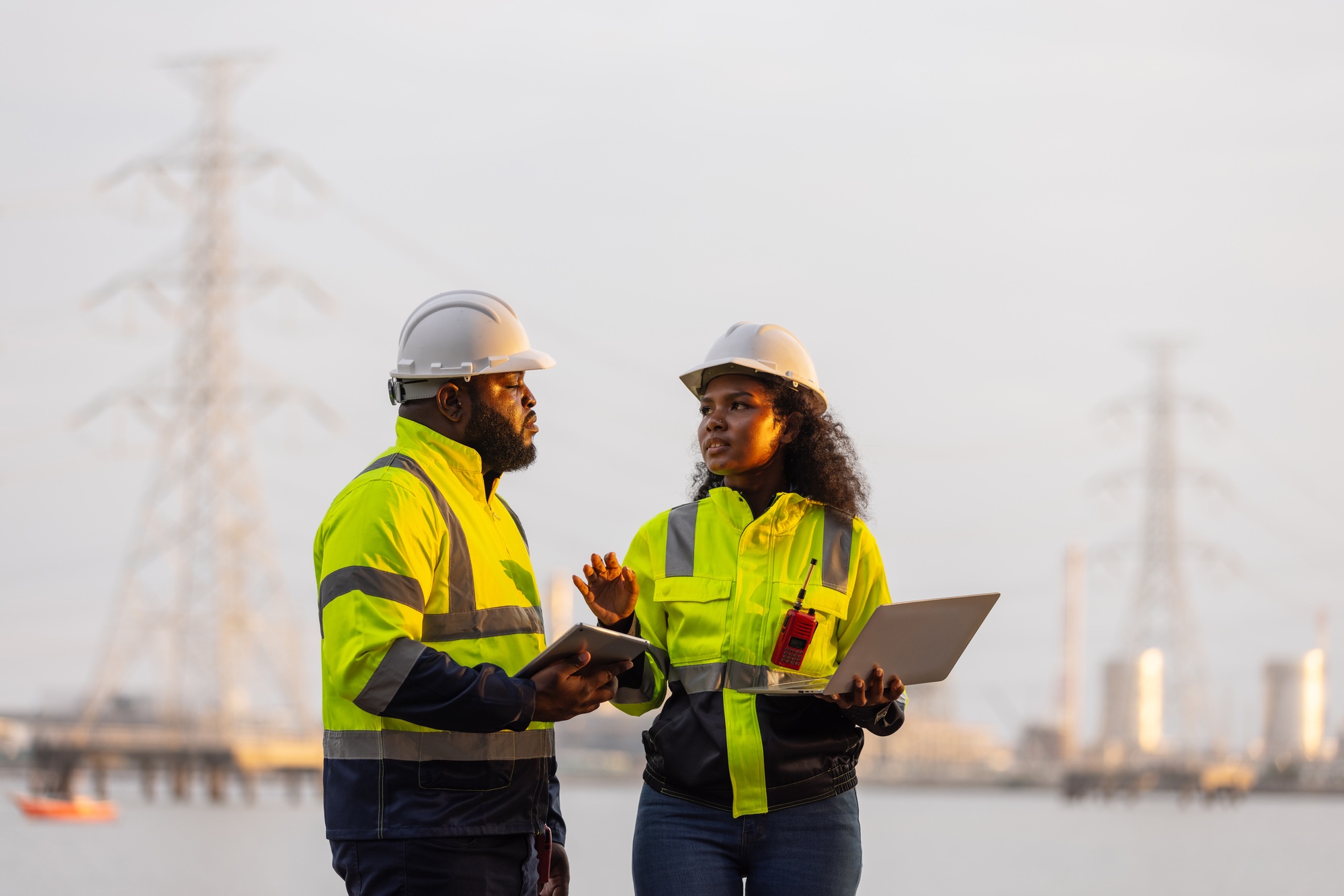 Two professionals in high-visibility jackets and hard hats stand in front of a power plant.